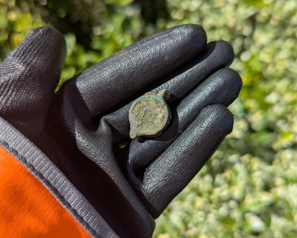 A gloved hand holds the Roman box seal. The circular box seal is around 2 centimetres in diameter and is tarnished green.