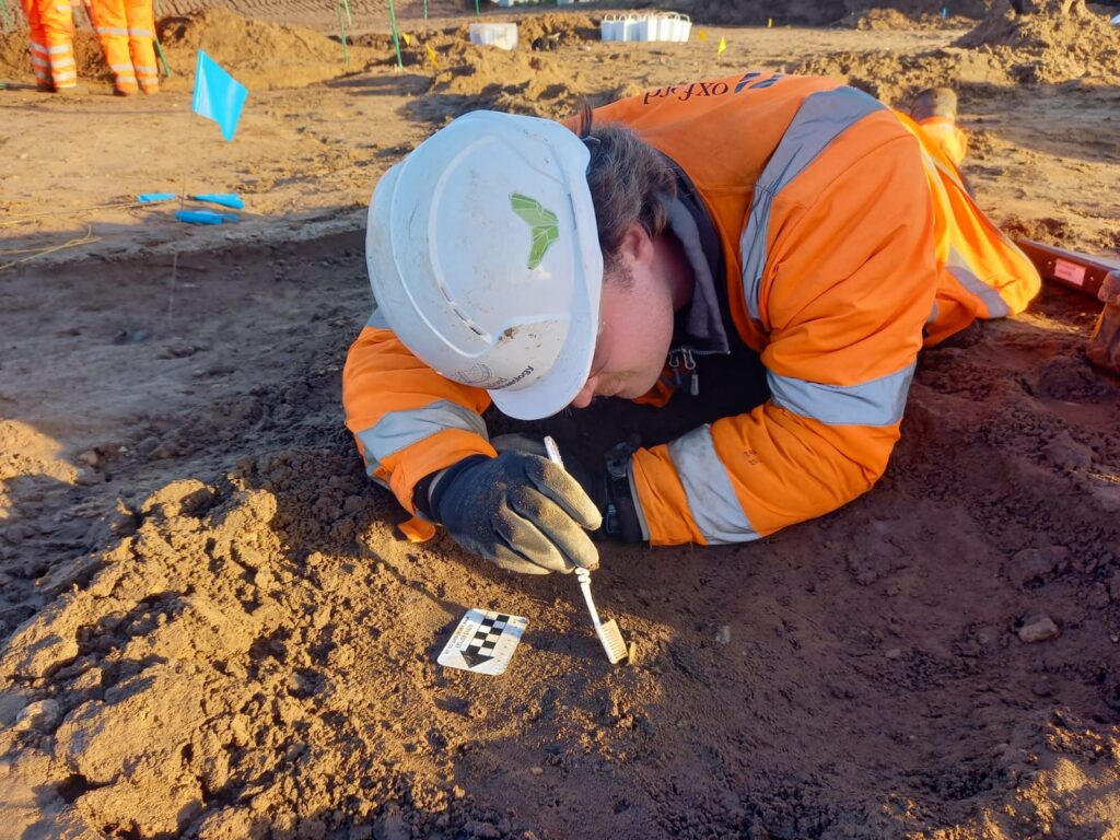 Hadley is lying on the ground facing the camera; he is looking down at a half-excavated coin in the ground which he is gently uncovering with a toothbrush