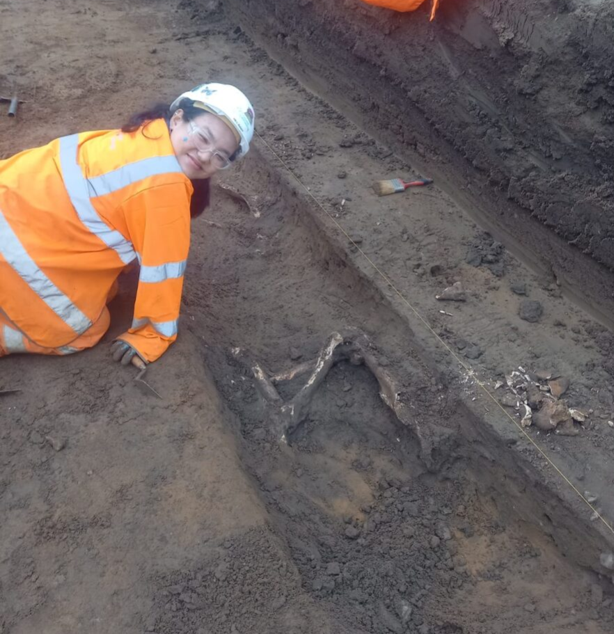 Megan is in the left of the frame, smiling at the camera and is kneeling to the the side of a partially excavated horse burial; some of the horse's leg bones are exposed.