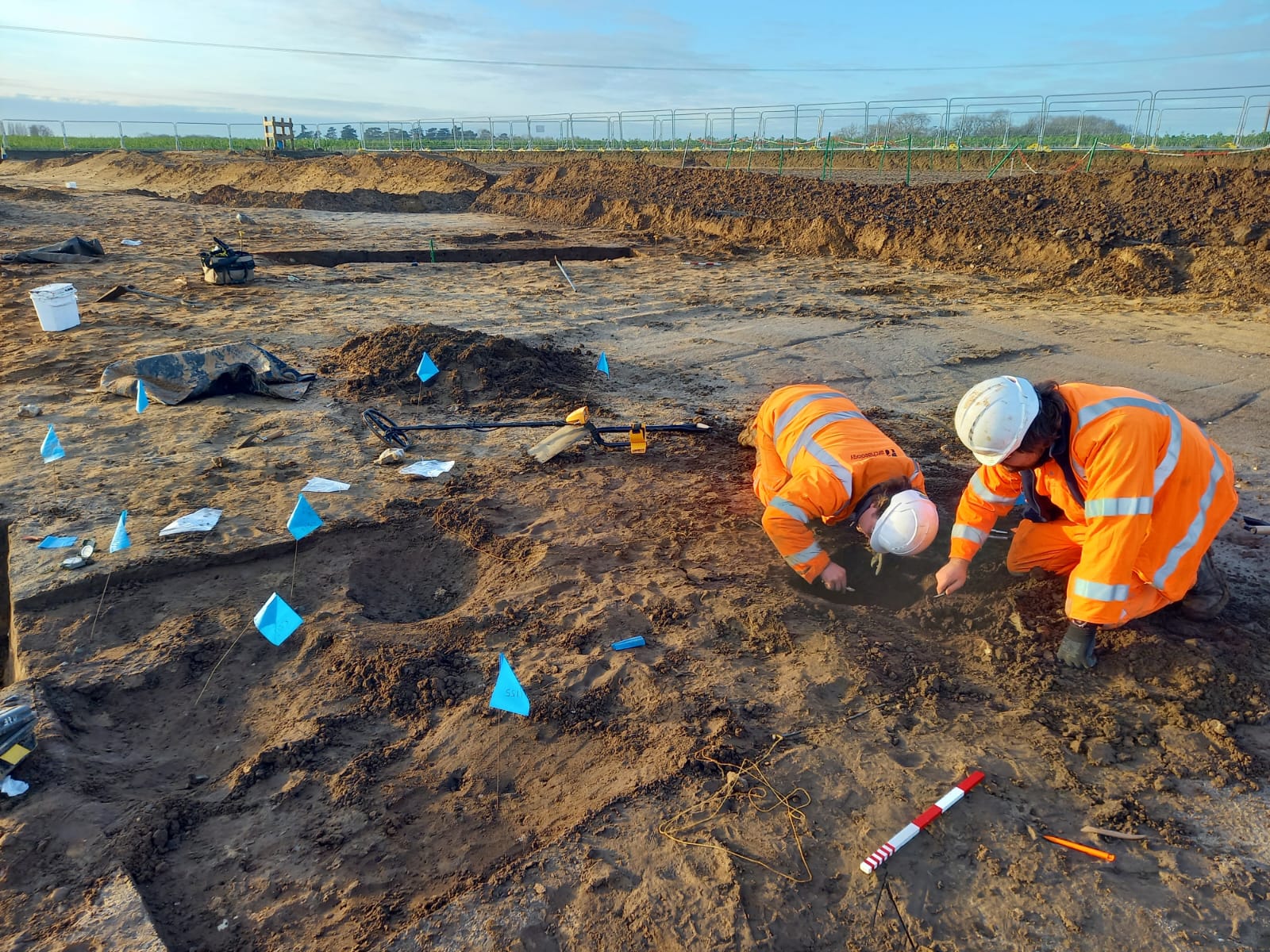 Two archaeologists are kneeling in the right of the frame. One is closely examining the feature they are both digging. The rest of the frame shows the stripped soil of the site with the horizon and blue sky in the upper third of the image