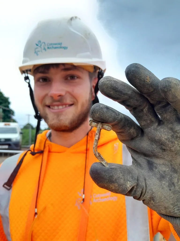 Smiling archaeologist holds a Roman brooch between their thumb and forefinger.