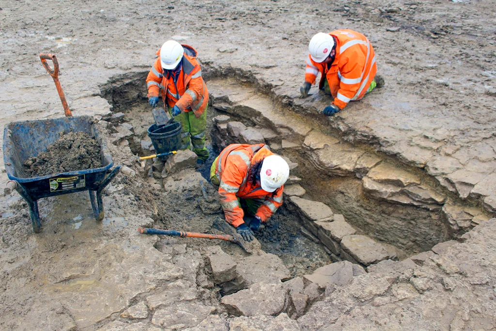 Three archaeologists in orange PEE excavating a stone lined corn dryer.
