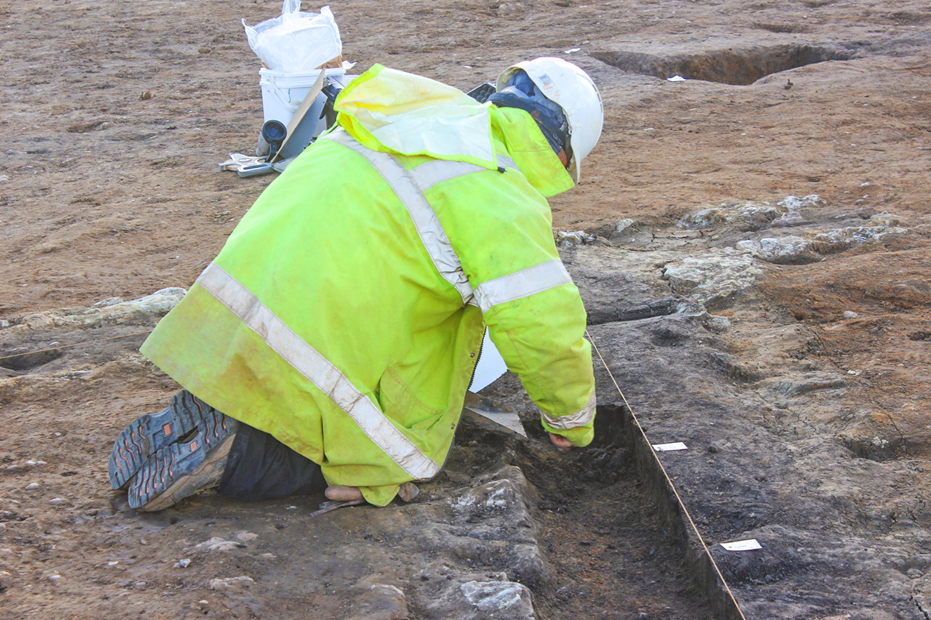 Photo of an archaeologists in green PPE excavating a slot through the flue of a Roman corn drier