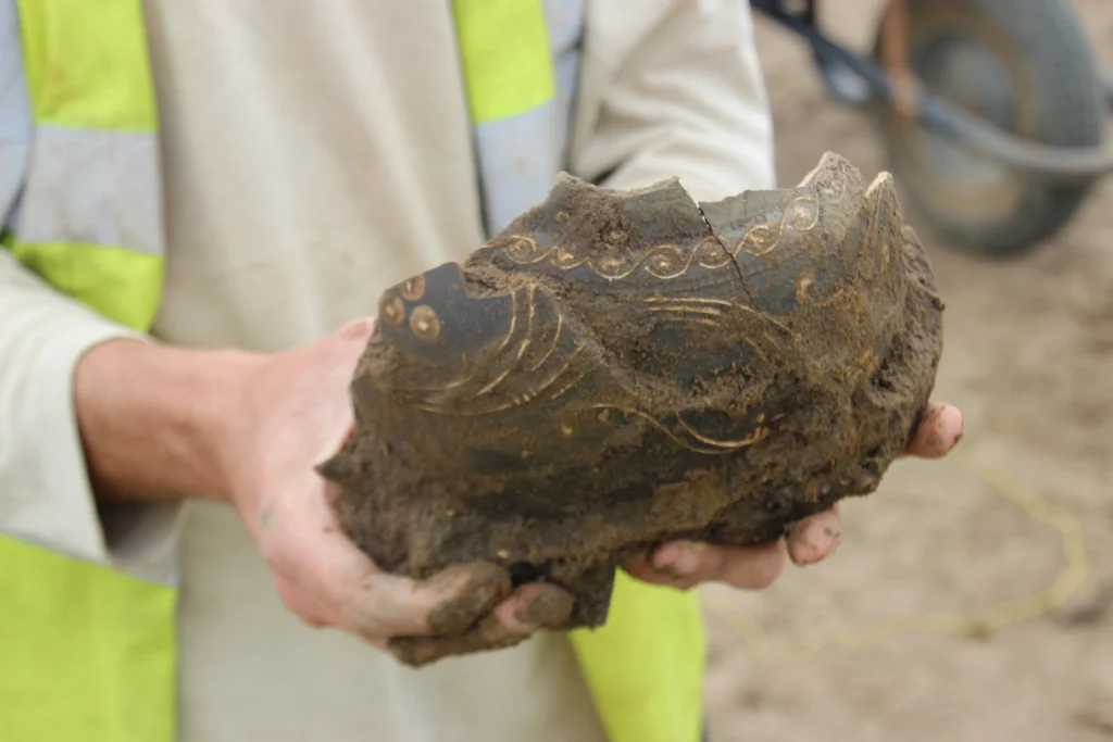 A nene valley pot with white decoration being held. It has been recently excavated and still has the mud which is holding the sherds together somewhat resembling its original shape