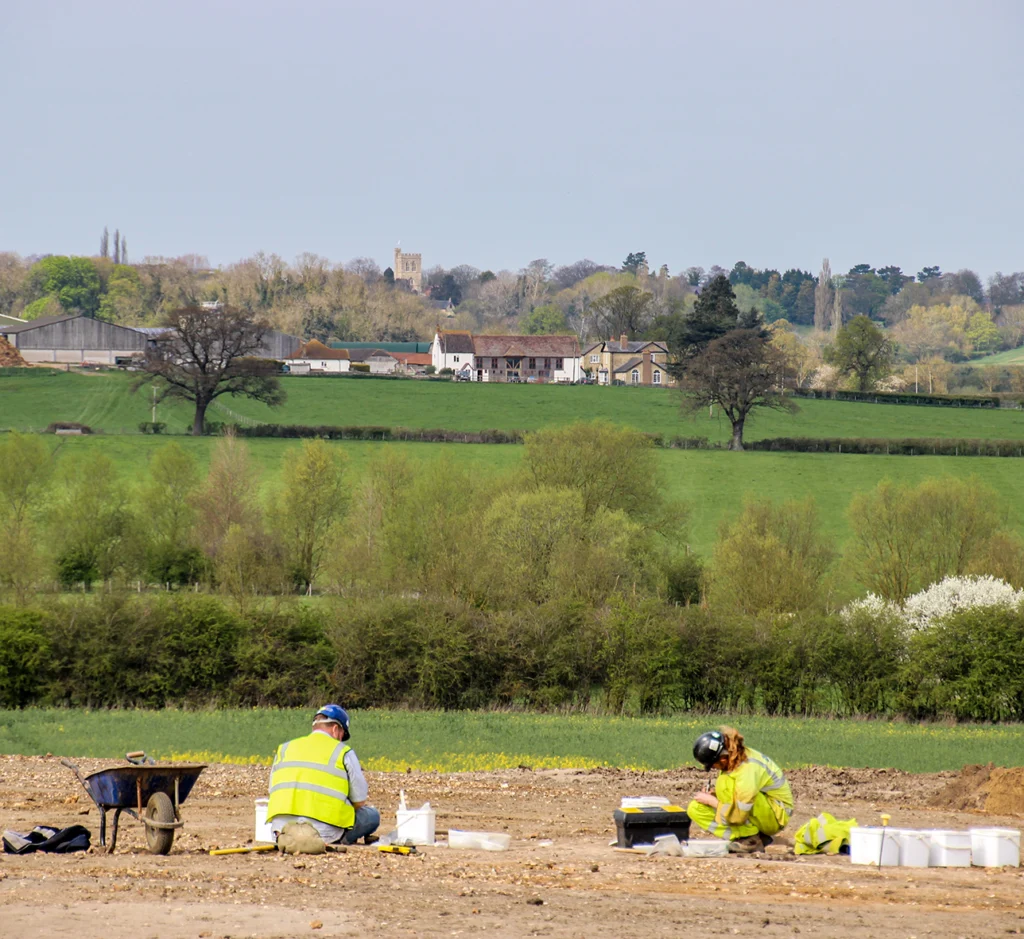 Archaeologists in yellow hi-vis protective clothing work on site in the foreground the a green landscape behind them