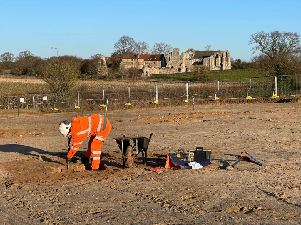 archaeologist in orange PPE excavating with a shovel next to a wheel barrow and tools, in the background there is a fence line for the excavations and behind that you can see the ruins of the Abbey's second site