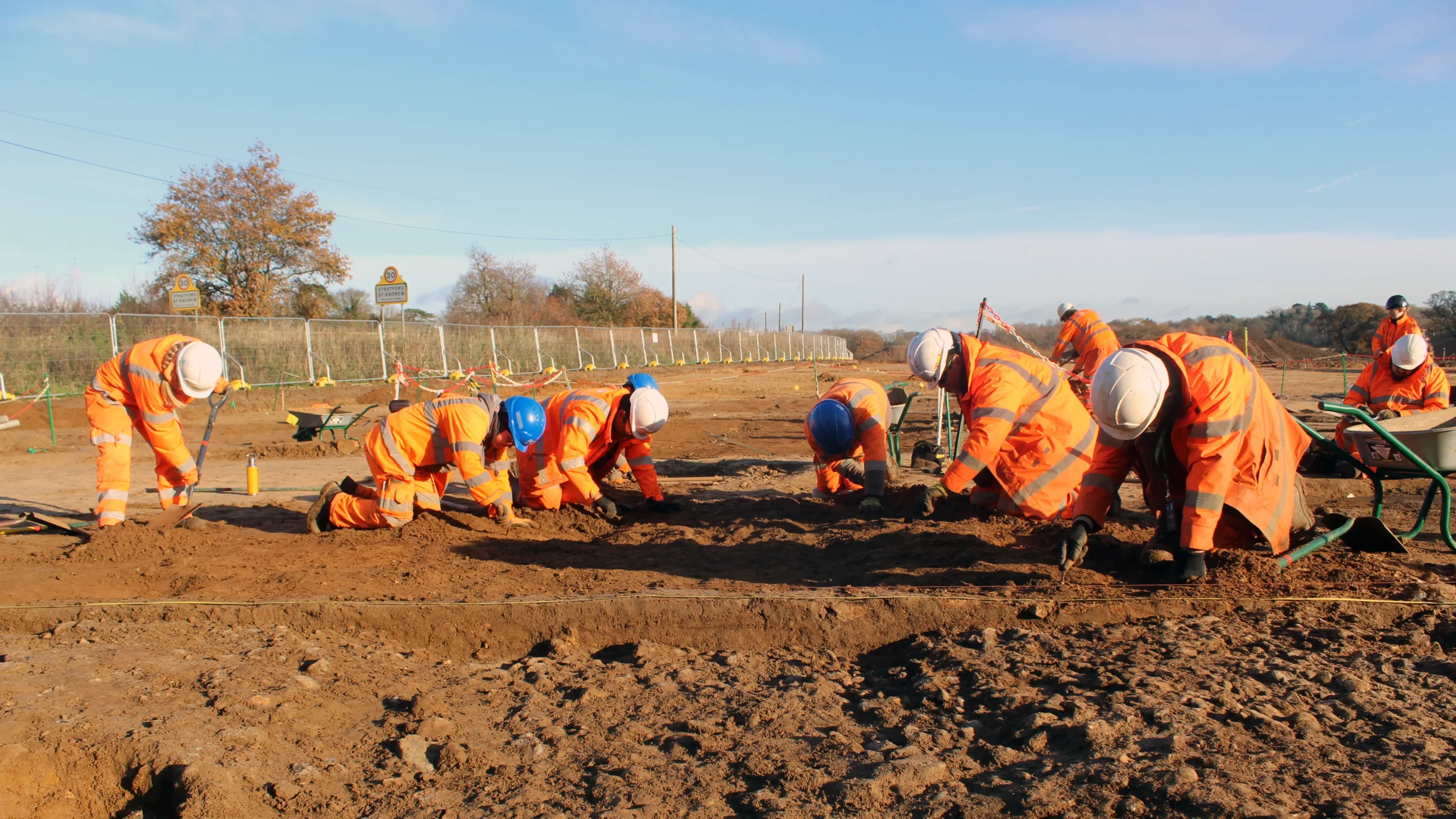 9 archaeologists all working on the TVB site with herris fencing running along the nearby road in the background with the village sign