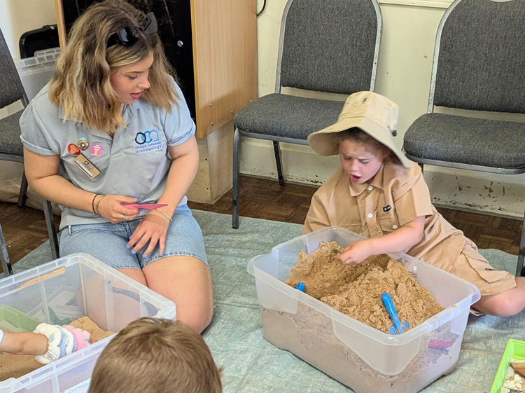 Photo of an OCA staff member sitting next to a sand pit activity looking surprised at a young girl who also looks surprised while she holds an item from a sand pit