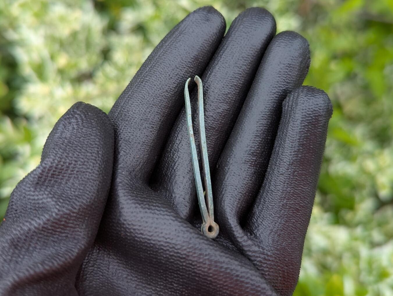 A gloved hand holds a pair of tweezers. They are tarnished green and the metal arms have a thick profile which tapers towards the ends.