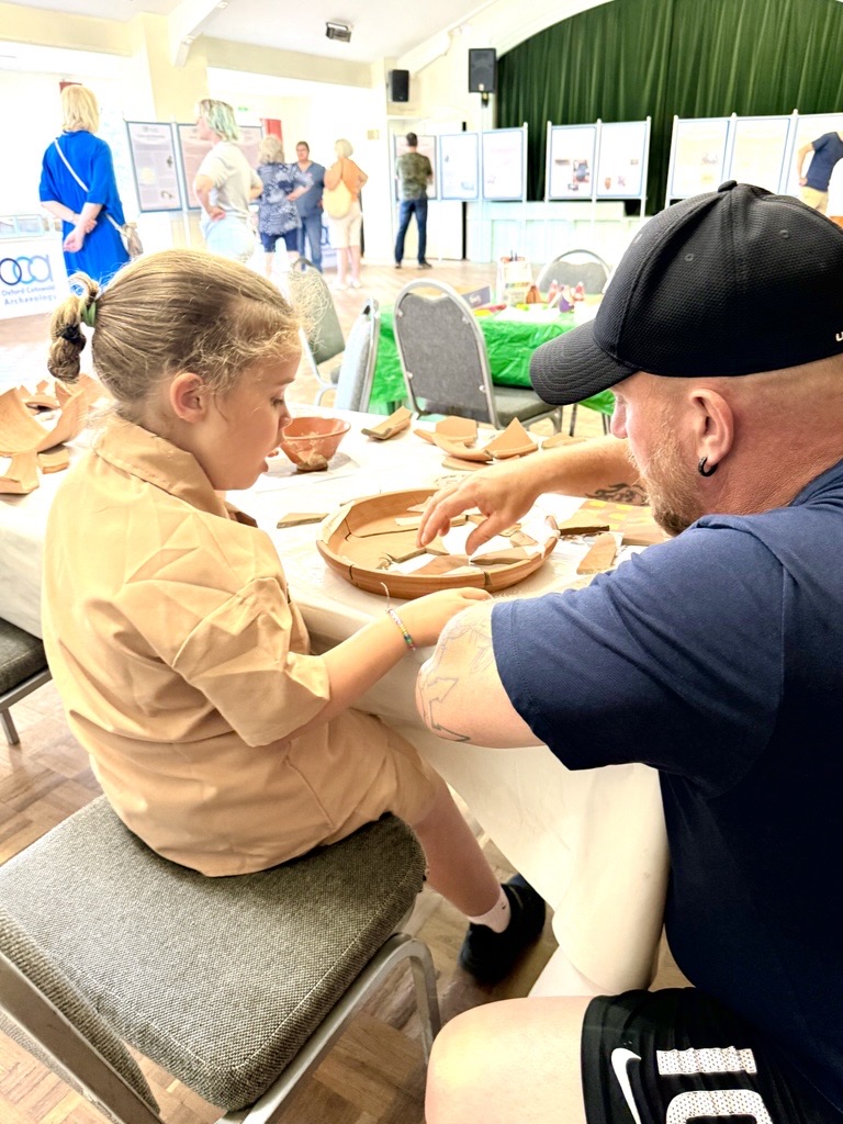 Photo of a young girl and her father sitting at a table and putting together pieces of a broken pot