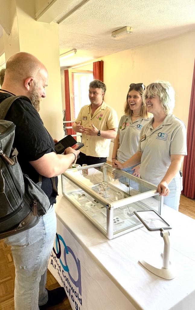 Photo of OCA staff members laughing with a visitor as he shows them photos on his phone in front of a display case