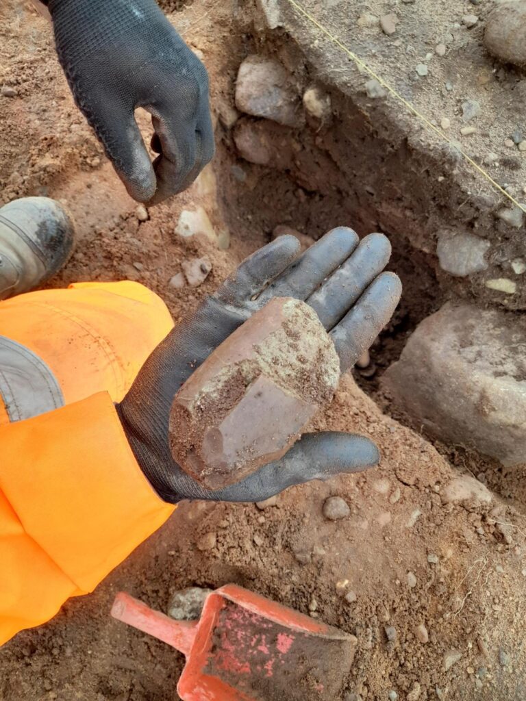 Archaologist holding stone axe in hands. Wearing gloves and orange PPE. 