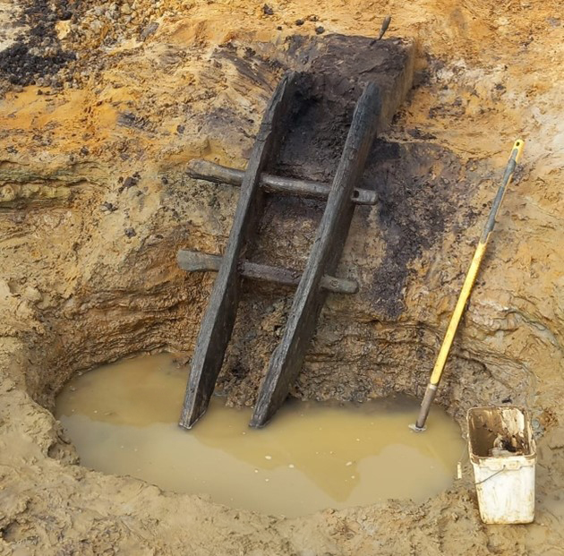 The wooden ladder with two remaining rungs sits partially excavated in a shallow pool of water