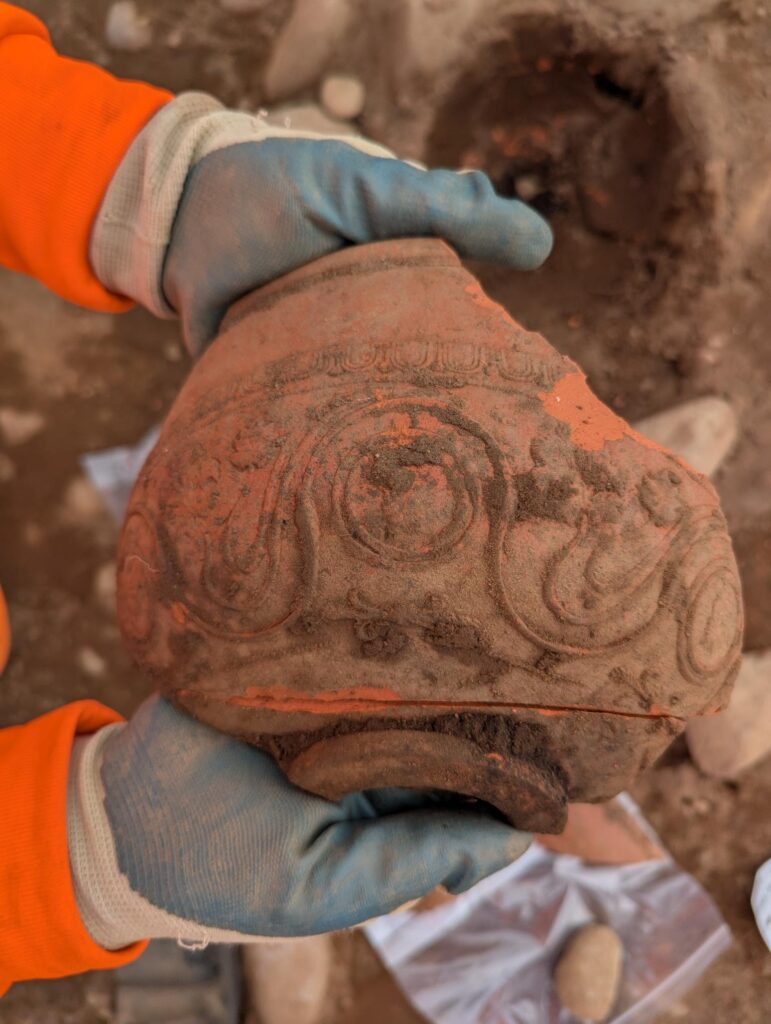 Archaeologist wearing blue gloves and orange ppe holding a large broken fragment of orange pottery. pottery decorated with swirls.