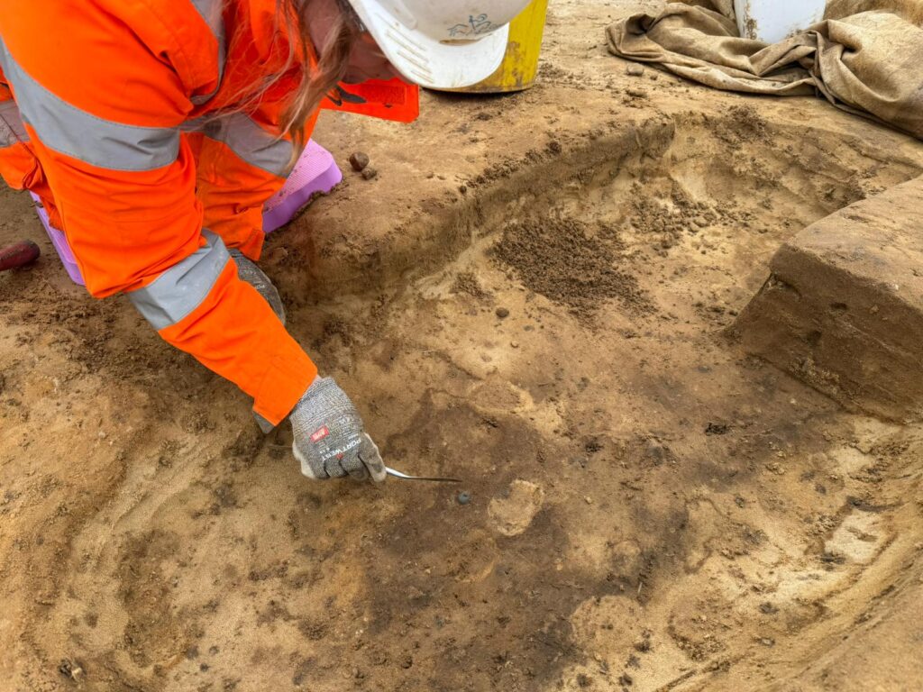 An archaeologist is kneeling over an excavated feature using a trowel. In the centre of the feature is a small blue Anglo-Saxon bead.