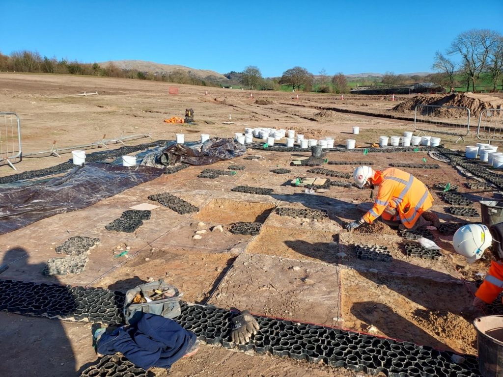 Landscape view of chequerboarding method with archaeologist in orange PPE kneeling down trowelling.