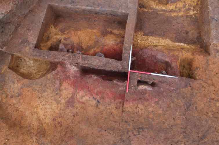 Rectangular archaeological excavation of a Roman saltern cut into yellow coloured soil. There are defined layers of red, brown and yellow and two 1 metre measuring scales in the centre arranged in a T shape.