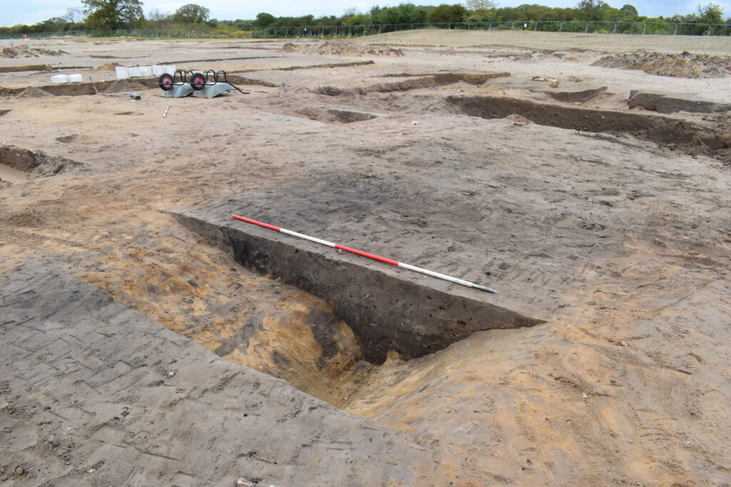An archaeological section of a dark brown ditch cut into yellow soils. There is a 2 metre scale rod placed on top of the ditch. There are two upturned wheelbarrows in the background.