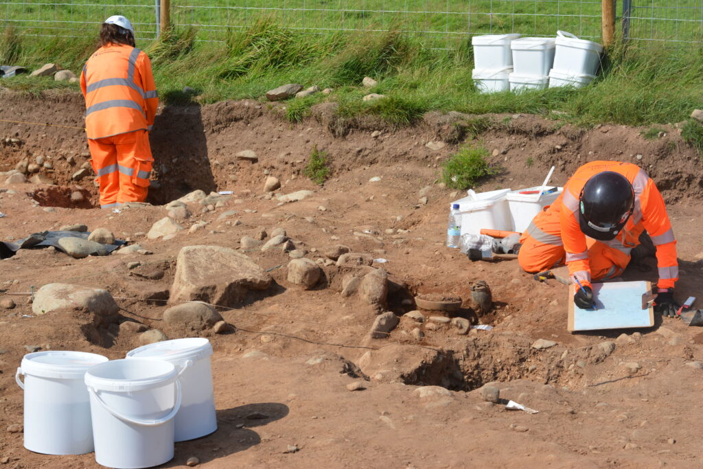 Archaeologist in front of cremation burial with visible vessels, holding a clipboard and a pencil. Surrounded by sample buckets.