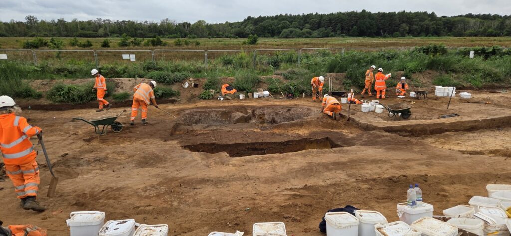 A team of archaeologists in orange high visibility clothing and white hard hats working across an open excavation, using trowels, hoes, and shovels. White buckets can be seen in the foreground, with a grassy area and trees visible behind the site.