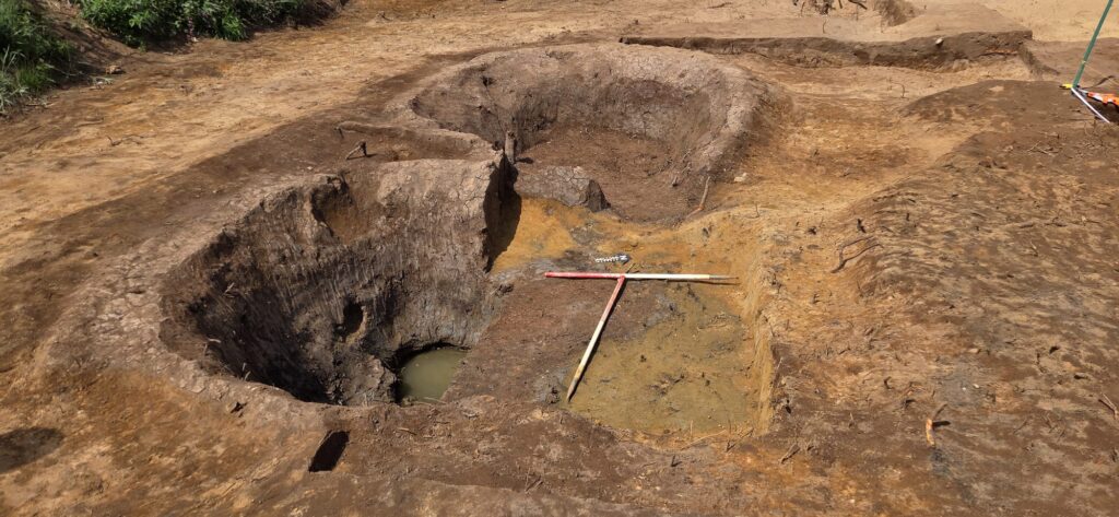 Two large circular archaeological pits which are the remains of Roman salterns. There are two 1 meter white and red scales positioned in a T shape in the centre of the image.