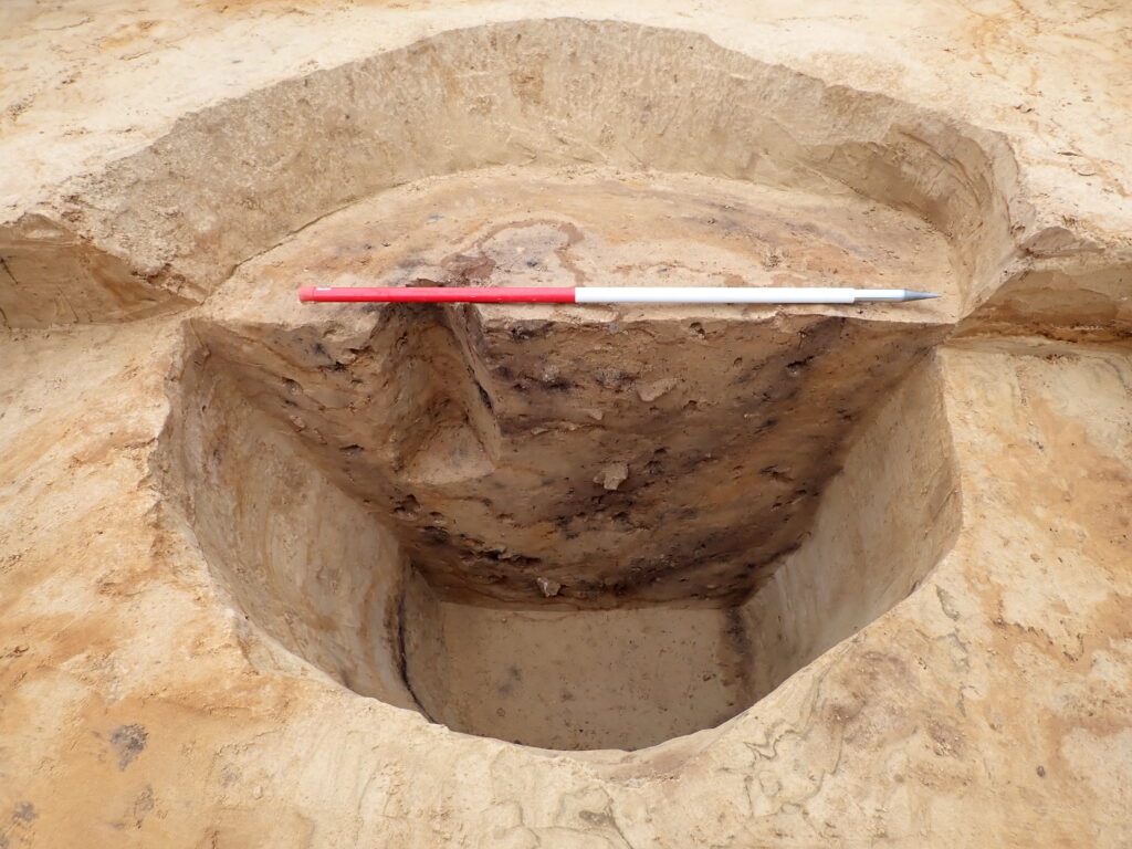 An archaeological half section of a circular pit measuring just over 1 m in both length and depth, showing clear layers of brown and grey soils.