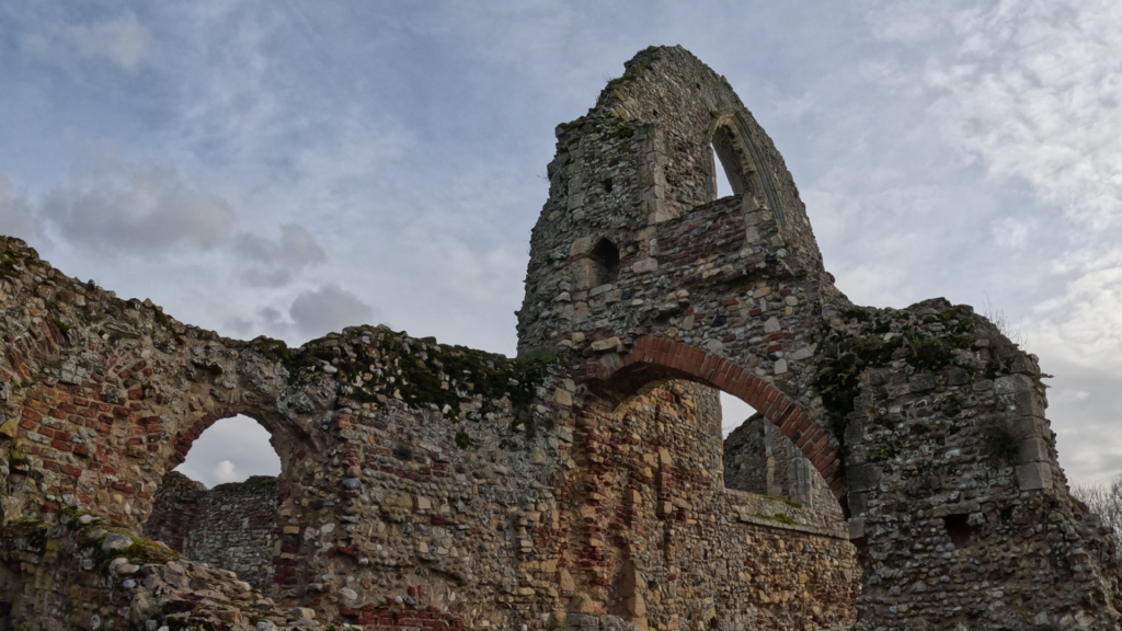 Stone ruins with partial walls and arches made from irregular masonry. Moss and vegetation cover some sections of the structure. The view is looking up at the ruins with a cloudy sky behind them.