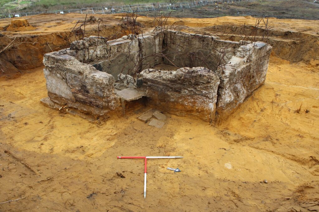 The remains of a square concrete structure with exposed metal rods protruding from the top of its walls. A T‑shaped measuring scale sits in front of it to indicate size.