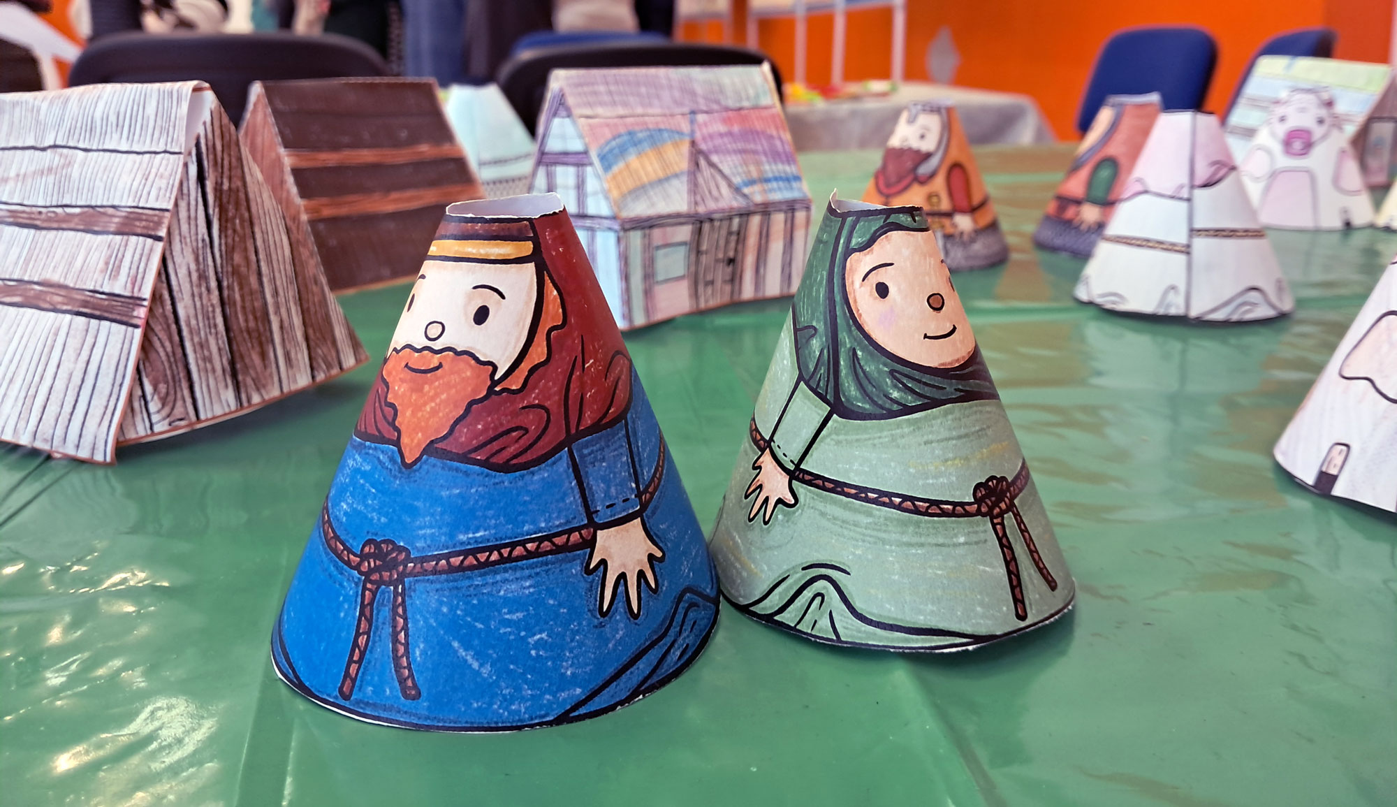 Close-up of two hand-drawn cone figures dressed in blue and green robes on a green table, with other similar figures and paper structures in the background.