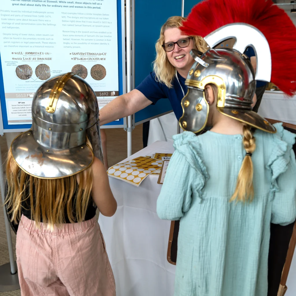 Two kids in replica helmets, the one in the left wearing the Sutton Hoo helmet, the one on the right wearing a Roman helmet, an OCA staff member looks into the camera and smiles