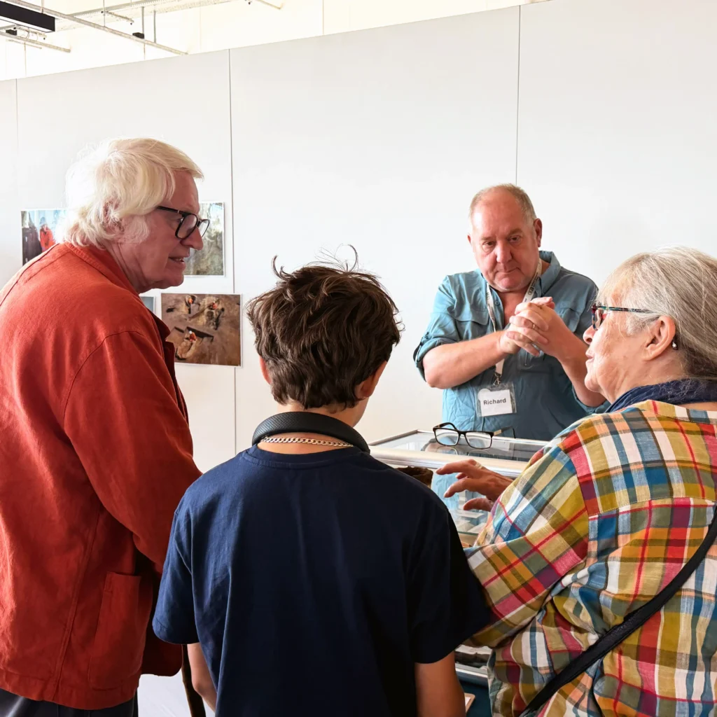 A group of four people, are gathered around a display case. The adult on the left wears an orange jacket and glasses, looking towards the display. In the background, a man with a name tag, is talking with his hands. To the right, an adult in a colorful plaid shirt leans in and gestures towards the case, speaking to the child in the foreground who faces away from the camera.