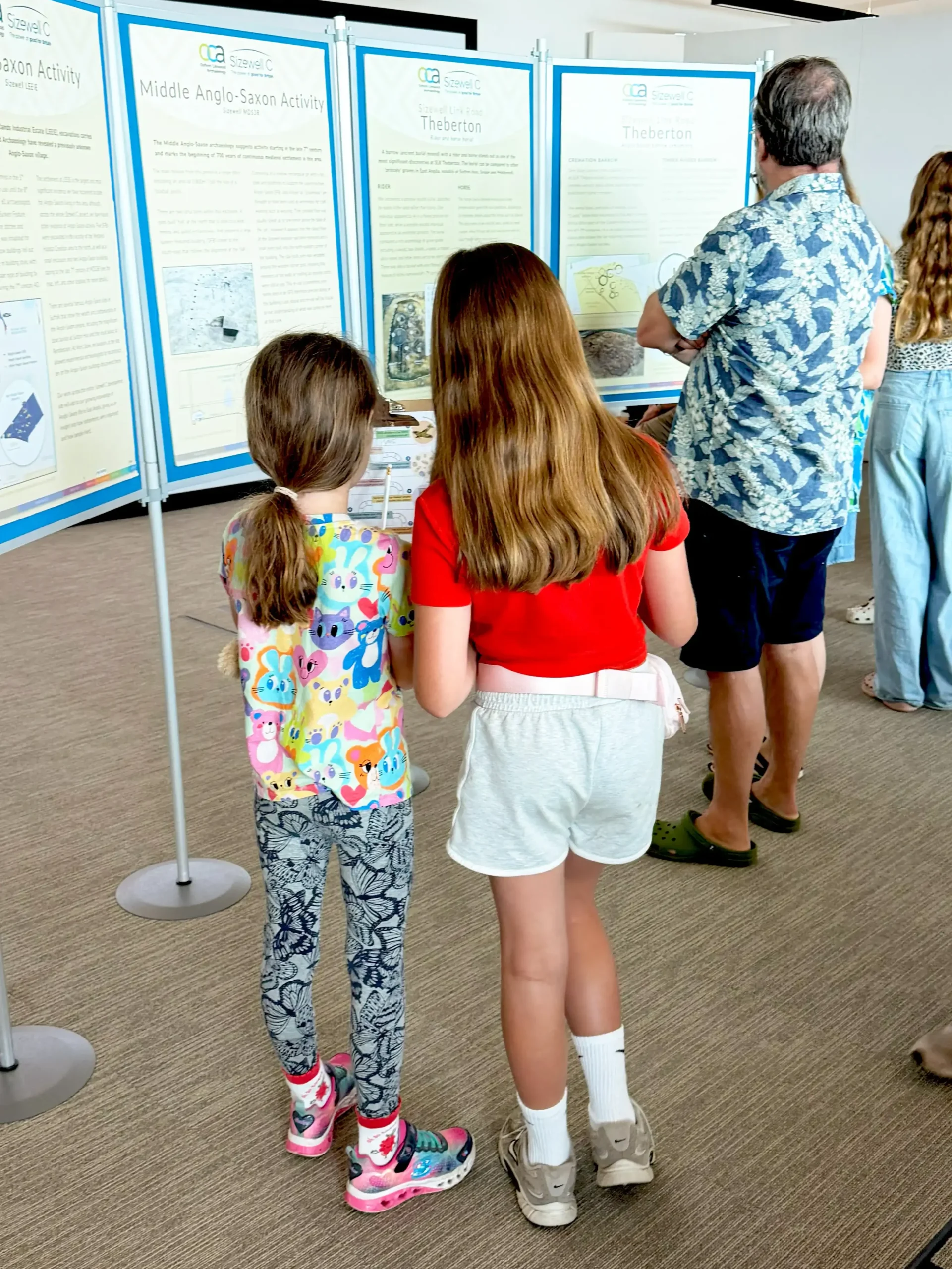 Two children stand with their backs to the camera, facing a display of information boards, one with a colorful patterned t-shirt and leggings, the other in a red shirt and gray shorts, with an adult in a blue patterned shirt and dark shorts also viewing the displays.