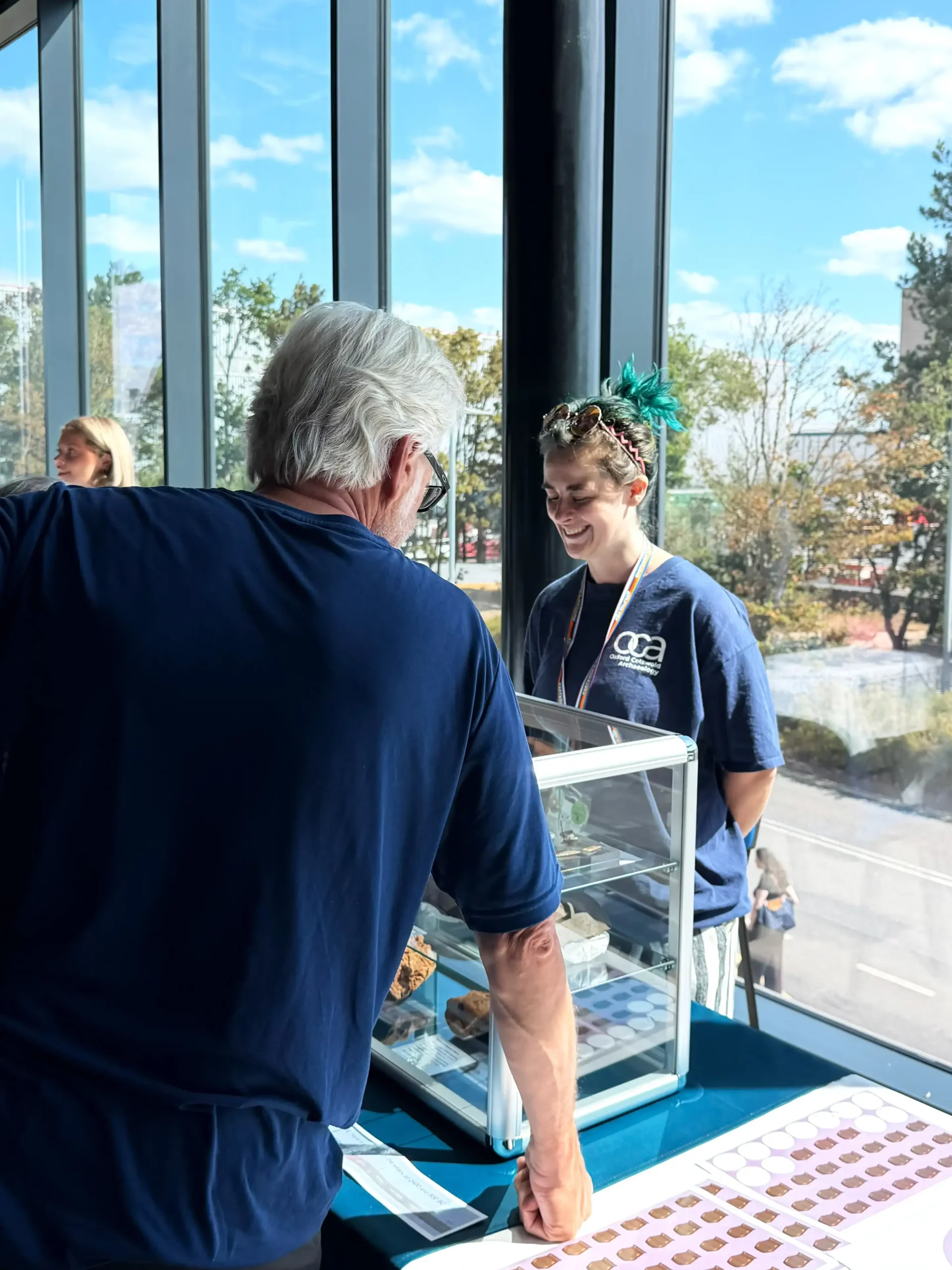 Two people interact at a table displaying artefacts in a glass case, with a large window overlooking a tree-lined street in the background.