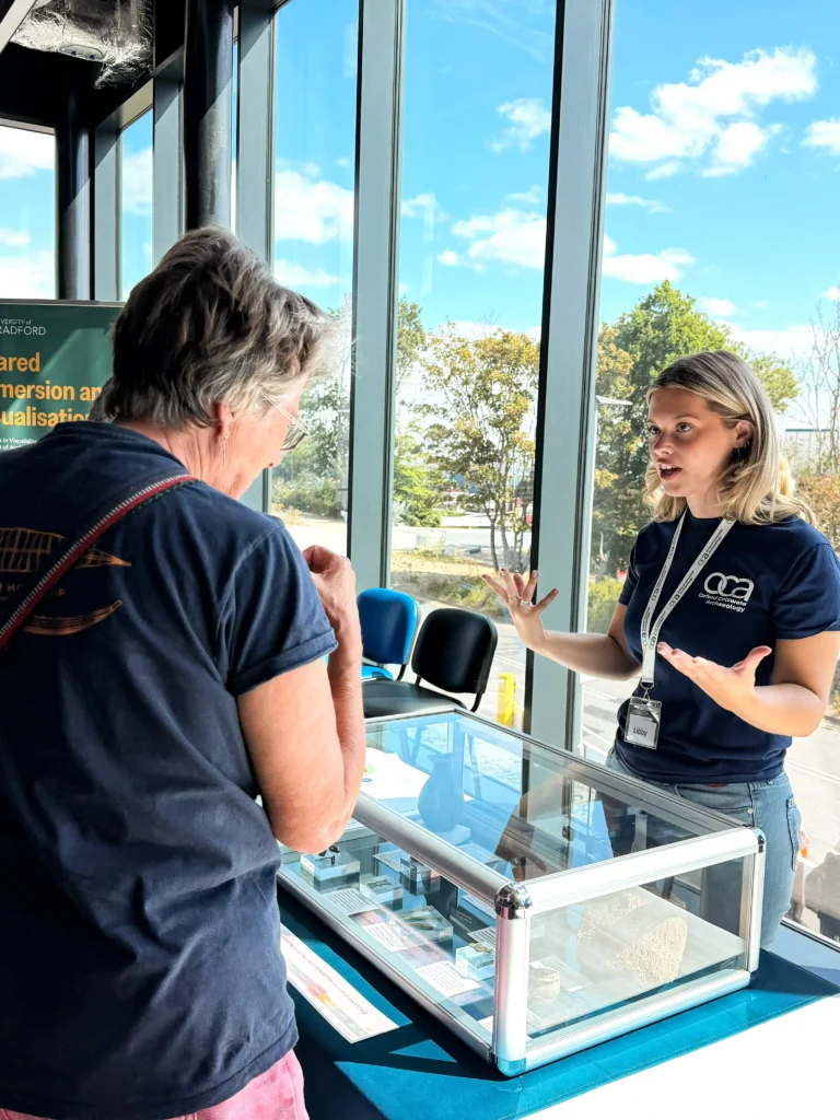 A woman in a navy blue t-shirt with an "oca Archaeology" logo is gesturing with her hands while speaking to a person in the foreground. They are standing at a display case filled with artifacts, in front of a large window with a view of trees and sky.