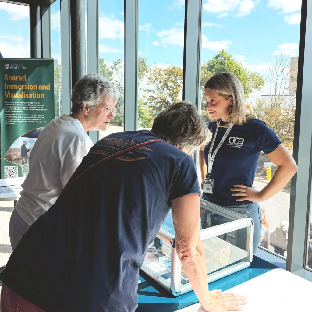 Three individuals are gathered around a display case in a bright sunlit room, with a banner for "University of Bradford Shared Immersion and Visualisation" visible in the background.