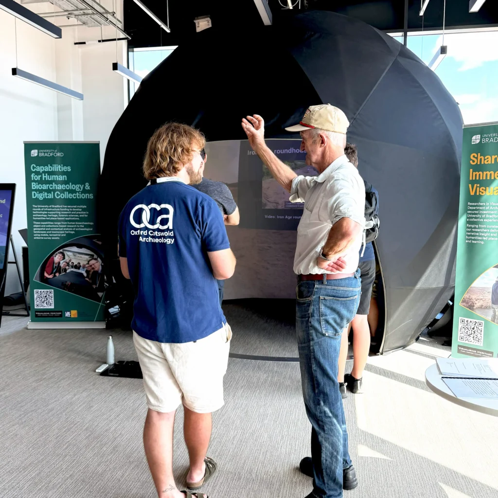 Two individuals interact near a large, black, dome-like projection screen displaying visuals of the excavations