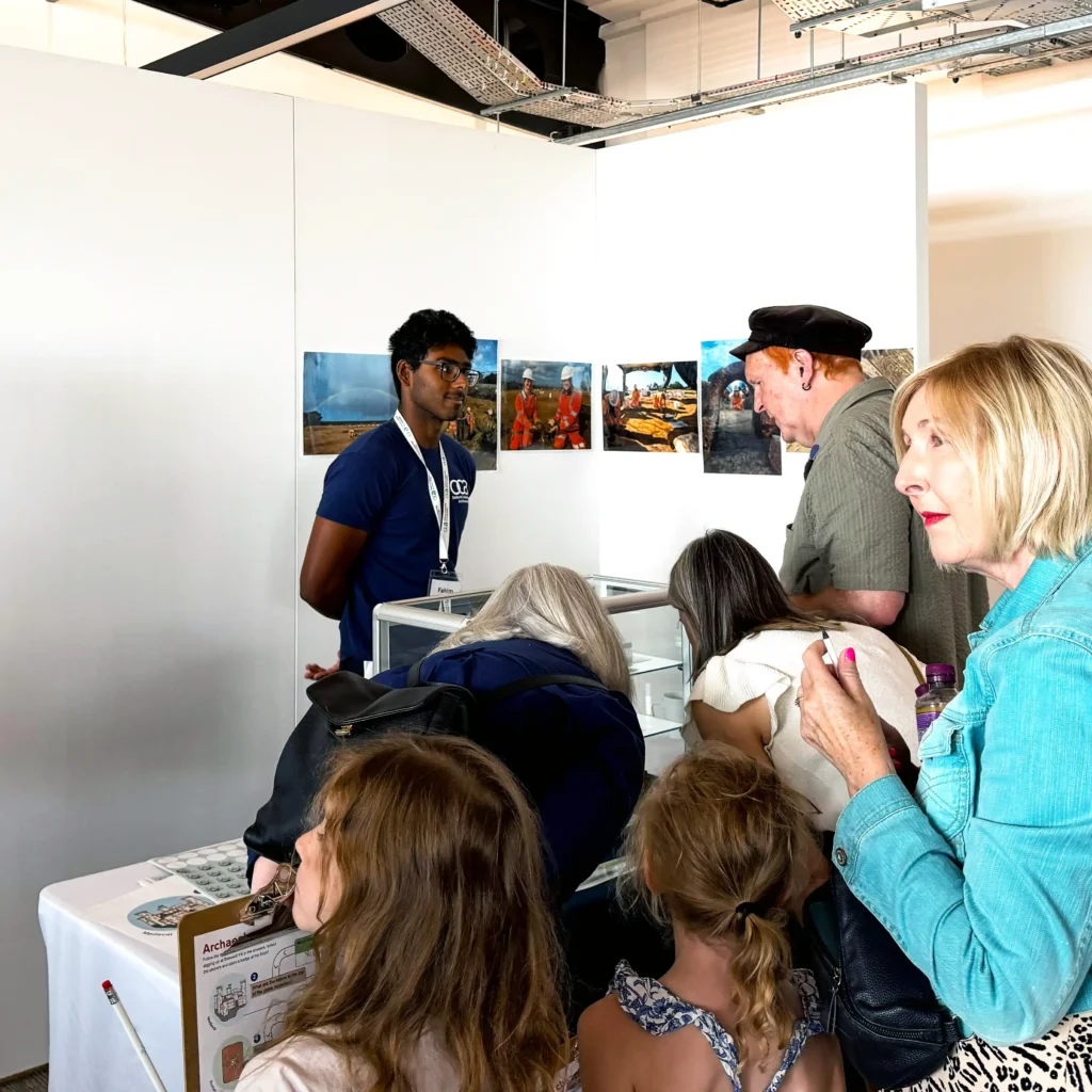 A man in a blue t-shirt explains something to a group of people gathered around a display case at an exhibition.