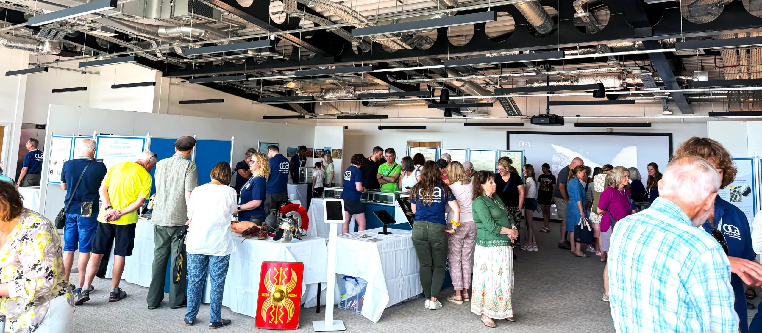 A group of people are gathered at tables displaying artifacts and information, at an archaeology exhibition, with a large projection screen in the background.