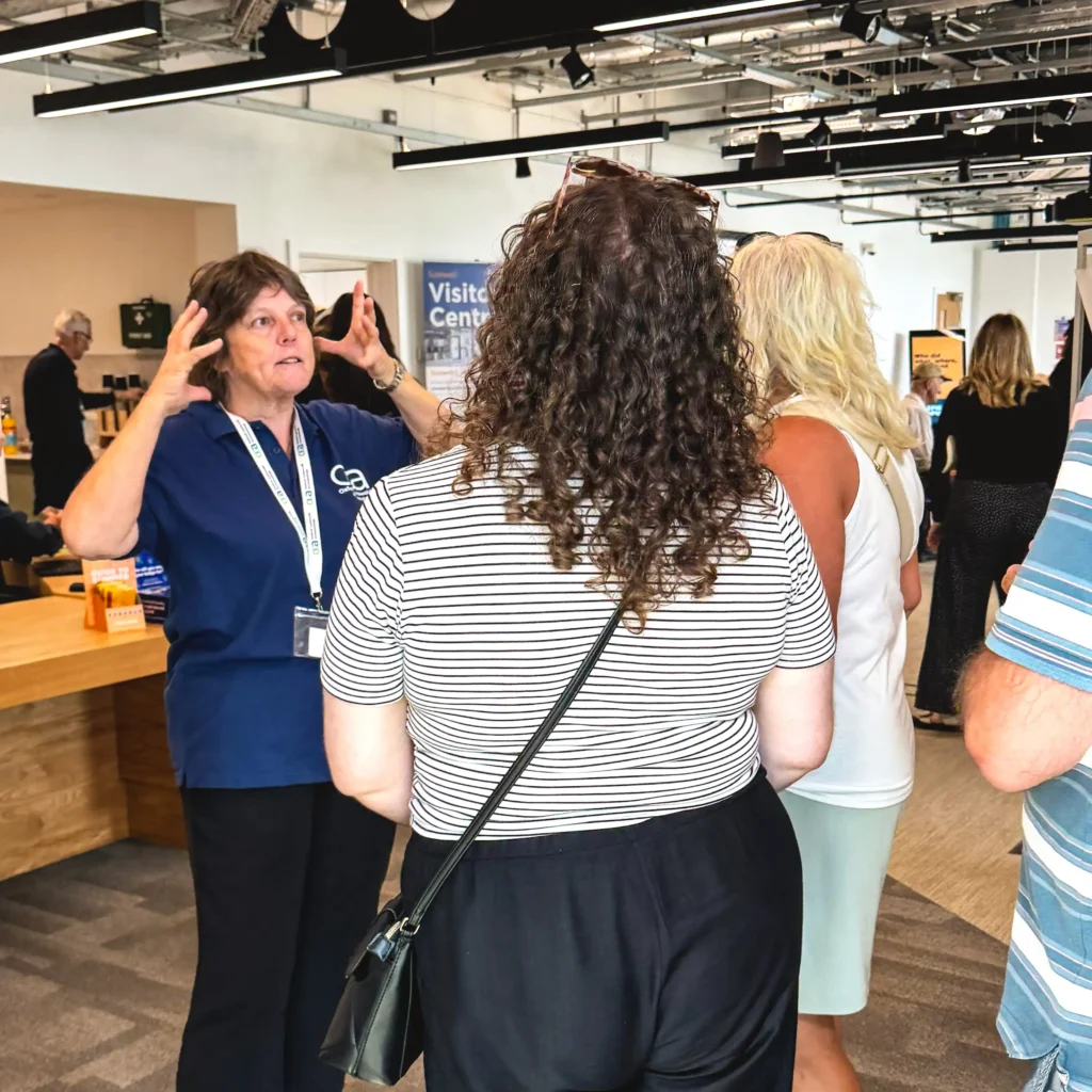 A woman in a blue polo shirt gestures with her hands while talking to a group of people in a bright, indoor setting with a "Visitior Centre" sign in the background.