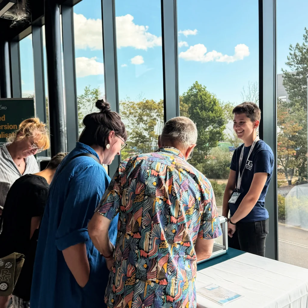 A group of people are gathered around a table, interacting with an OCA staff member wearing a dark blue t-shirt and lanyard. The background features large windows with a view of trees and a bright blue sky with clouds.
