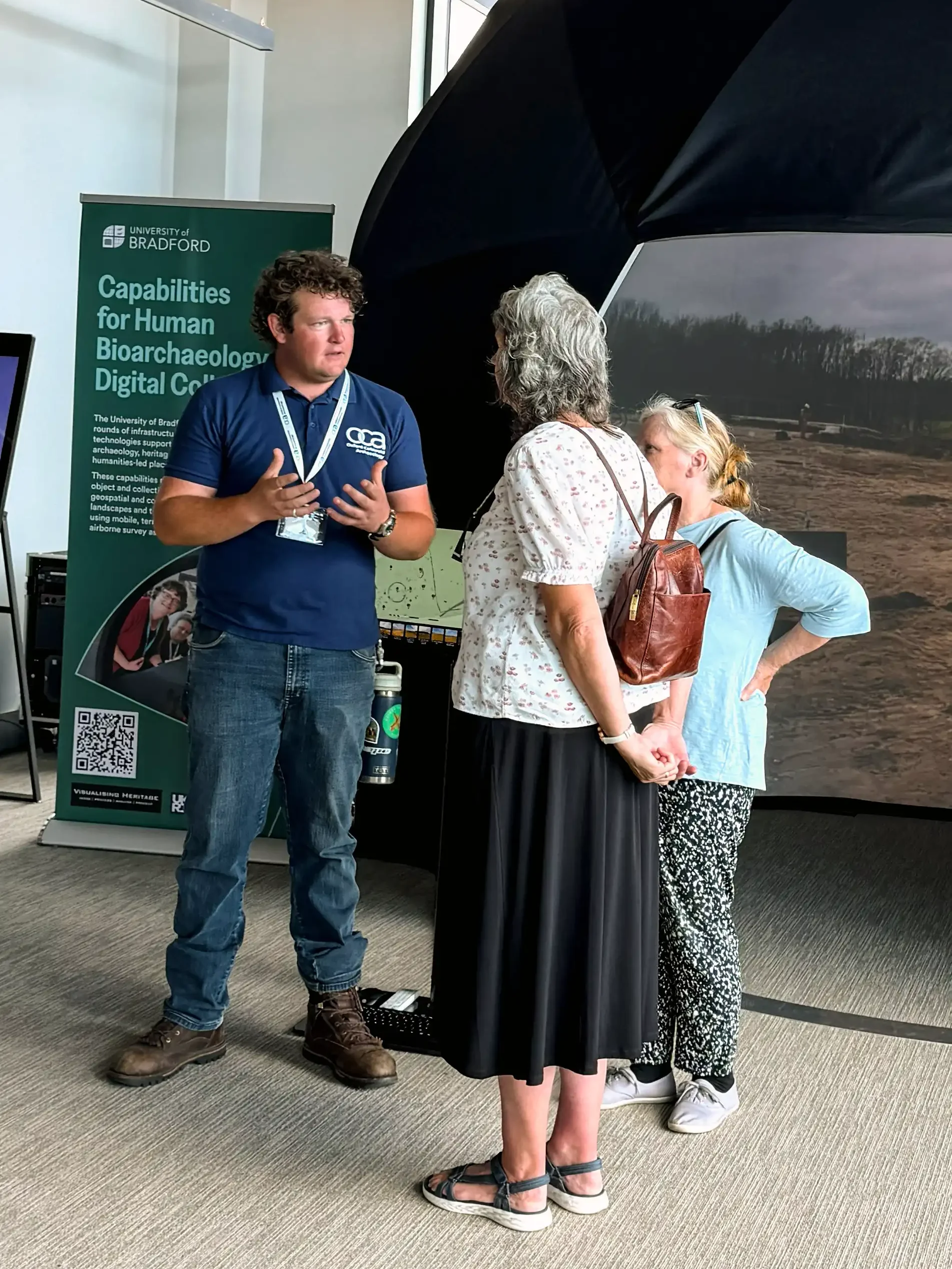 A man in a dark blue polo shirt and jeans gestures with his hands while speaking to two women at a University of Bradford digital archaeology exhibit.