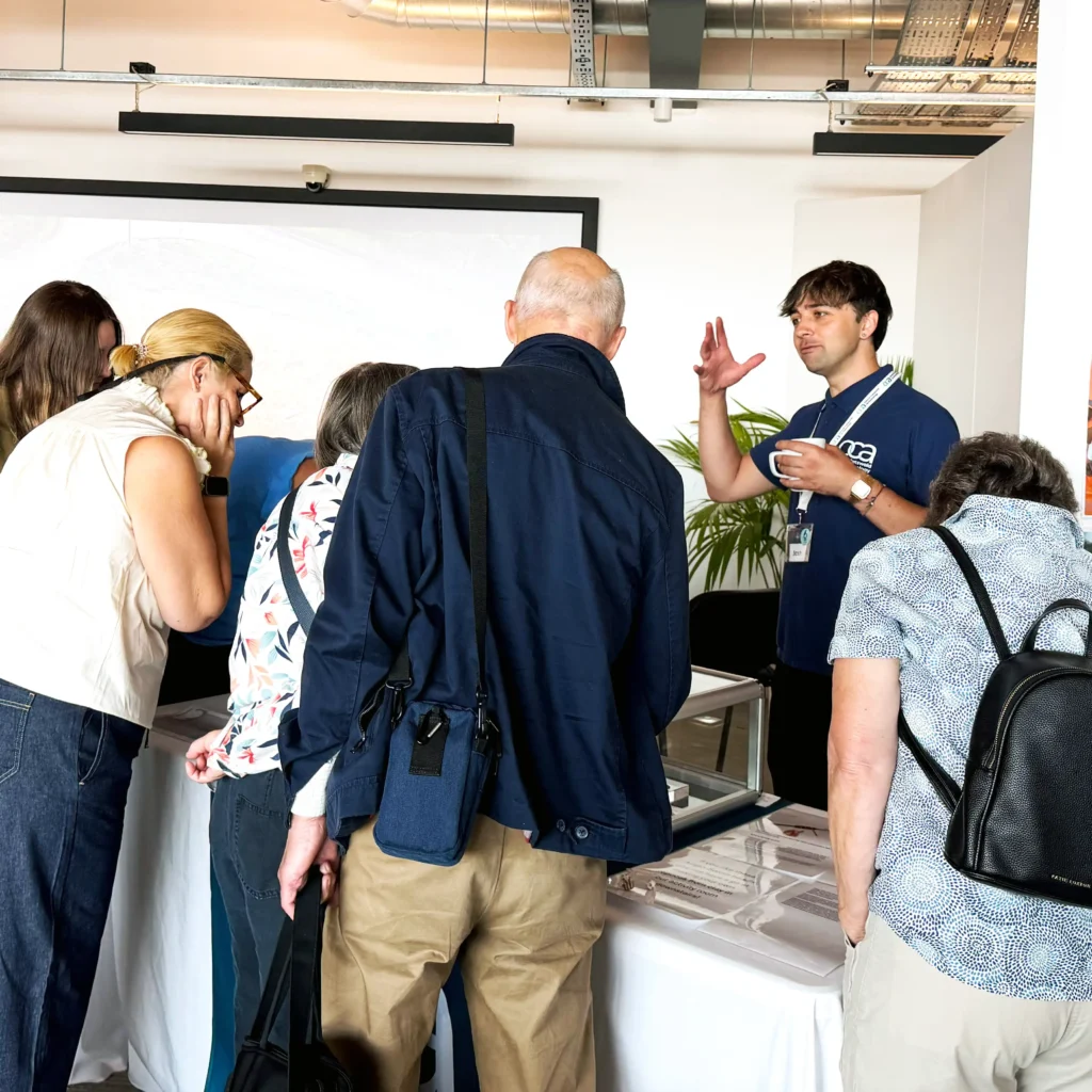 A presenter in a blue t-shirt stands next to a display table, gesturing with his hands while holding a cup, as a group of people lean in to examine the items on the table.