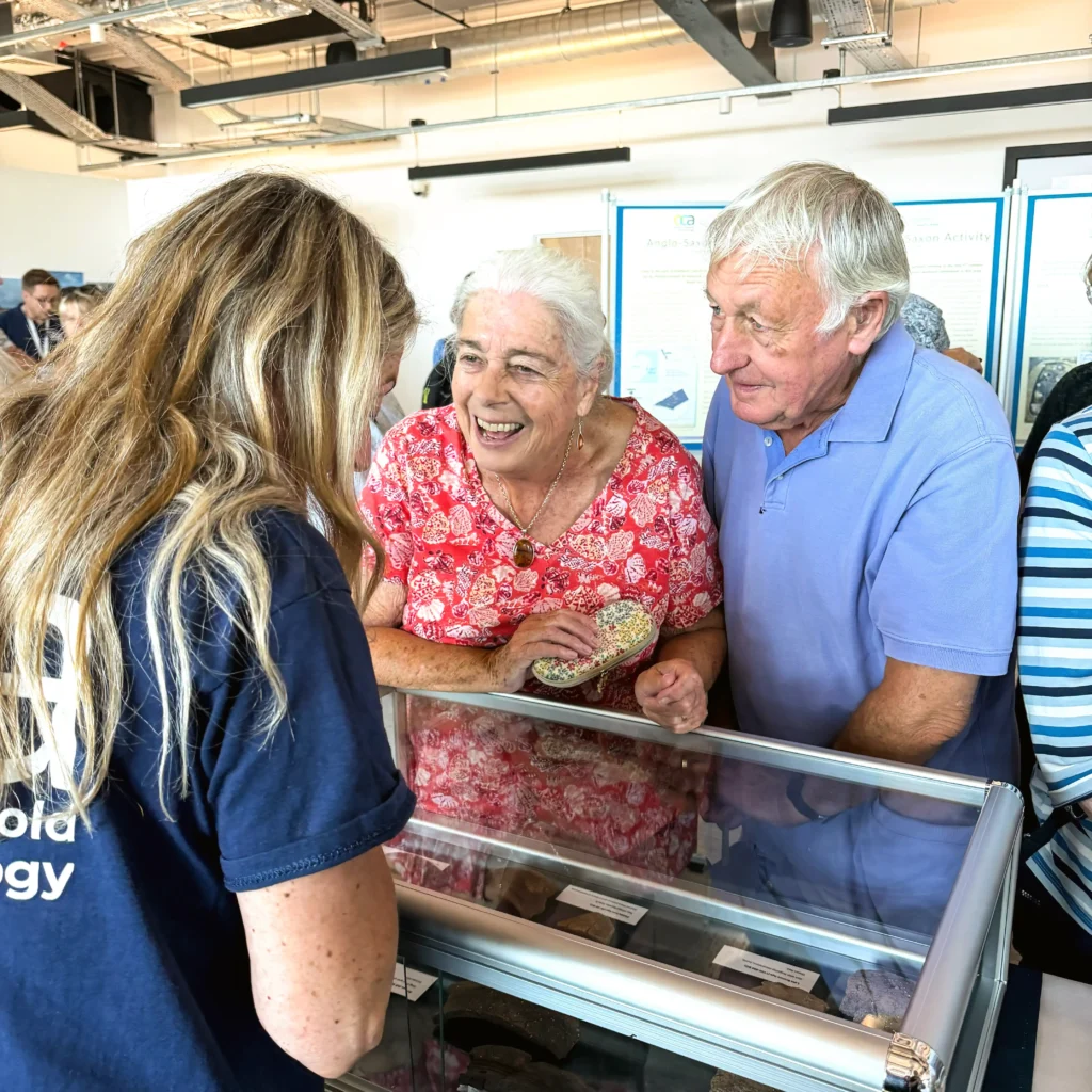 An older couple excitedly looks at artefacts displayed in a glass case at an exhibition, with a presenter in a dark blue t-shirt visible on the left.