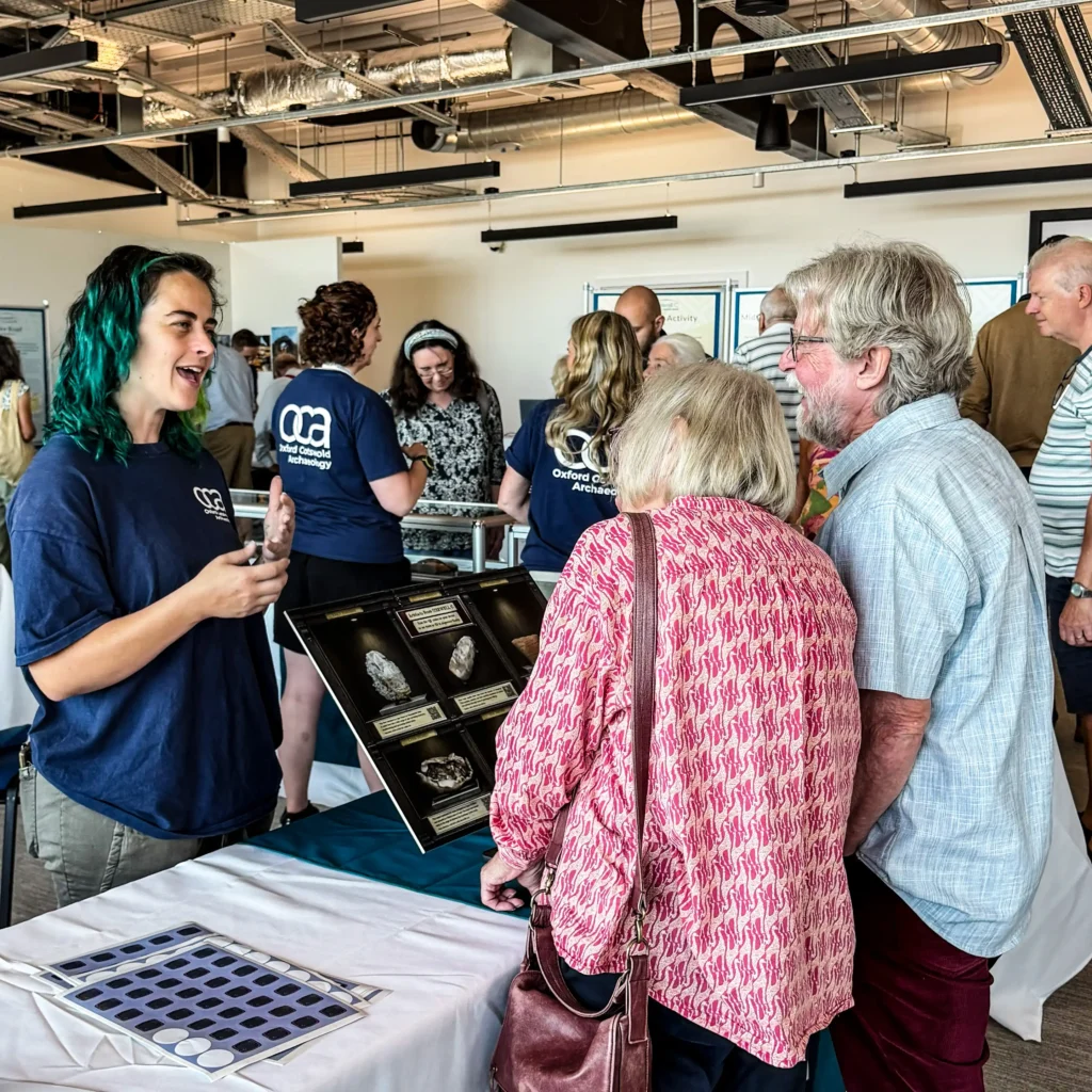 A group of people gather around a display table featuring archaeological artifacts at an exhibition. A woman with bright green hair, wearing a navy blue shirt with an "OCA" logo, gestures with her hands as she speaks. In front of her, a display boards shows several objects. Other visitors, including a man with a grey beard wearing a light blue plaid shirt and a woman in a pink patterned top, are looking at the exhibit.