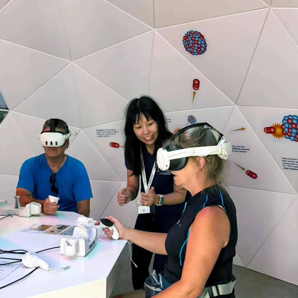 Two adults wearing VR headsets interact with a virtual reality simulation of site while a third person stands behind them, looking on and gesturing.