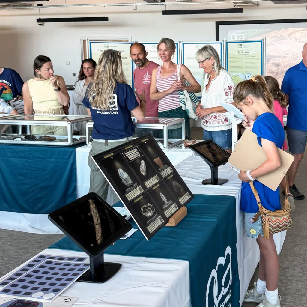 An archaeology exhibition with several display tables featuring artifacts and informational posters. Several adults and children are gathered around the tables, looking at the exhibits. One woman in a blue t-shirt is speaking to a group of visitors.