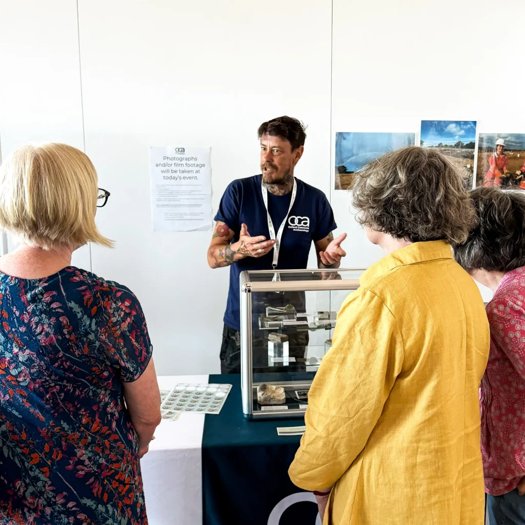 A OCA staff member in a dark blue t-shirt with tattoos gestures while speaking to two attendees at a display table featuring a glass case with artifacts.