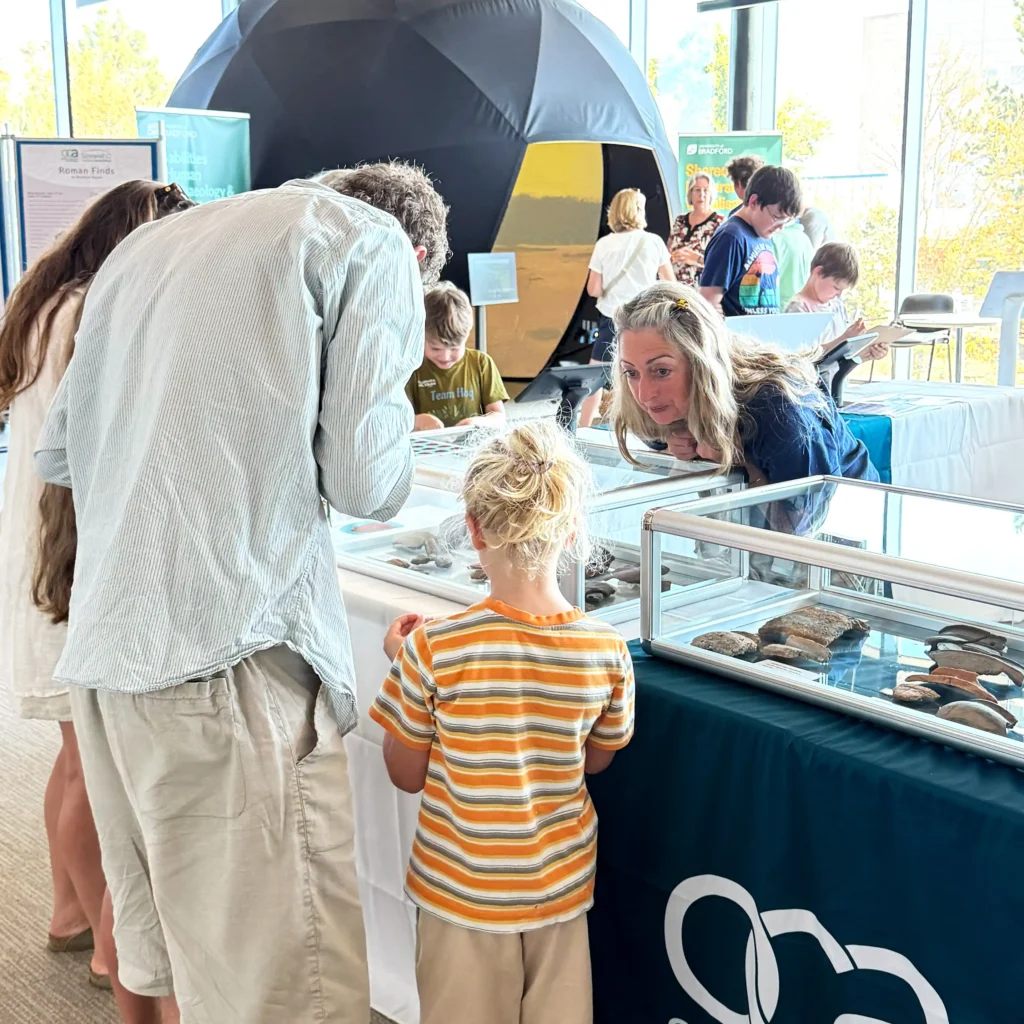 A group of people, including children, are gathered around display tables at an exhibition. The tables showcase artifacts in glass cases. In the background, a large black geodesic dome structure is partially visible, along with informative displays and other exhibition displays.