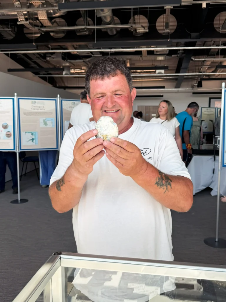 A man in a white t-shirt examines a large, rough, white and grey flint at an exhibition with informational posters in the background.