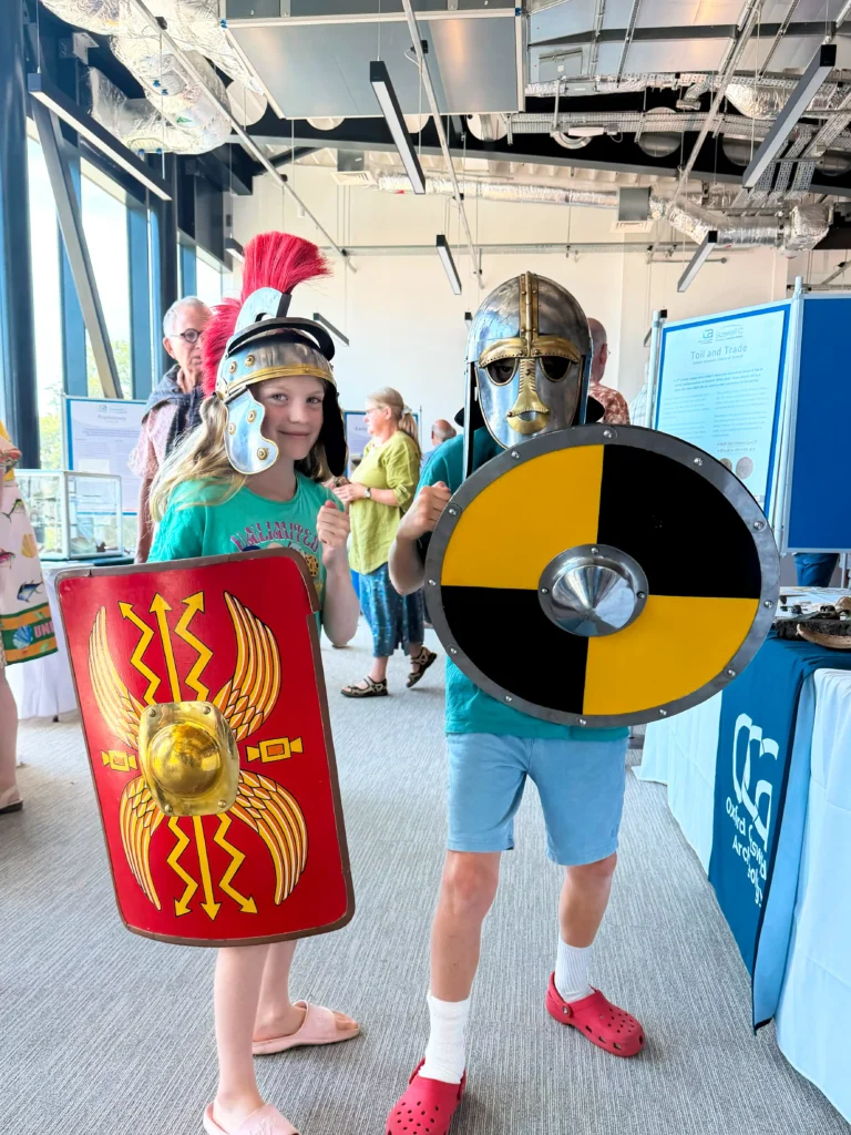 Two children dressed in historical warrior costumes, one with a Roman-style helmet and shield, the other with a Anglo-Saxon-style helmet and a round shield.